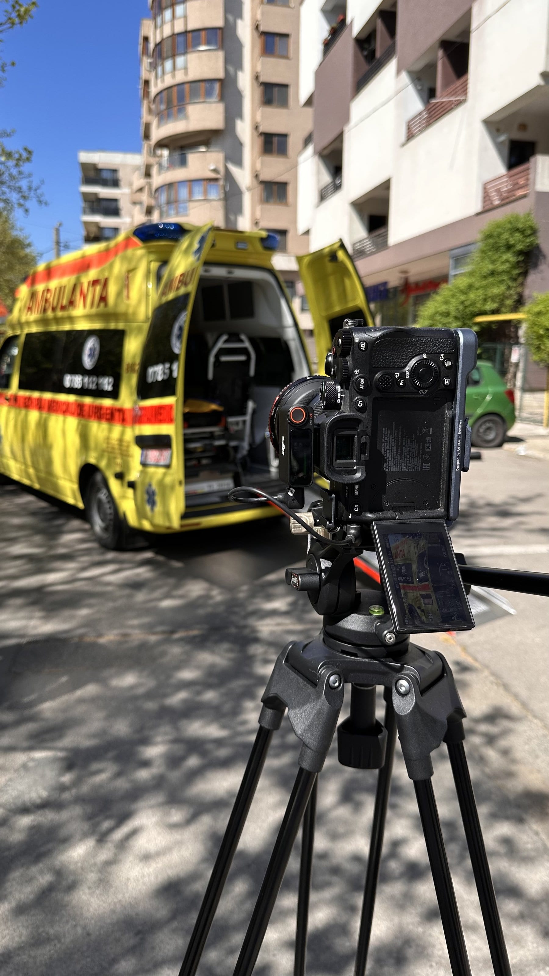 Cameră video pe trepied, filmând o ambulanță cu ușile deschise, pe o stradă din cartier.