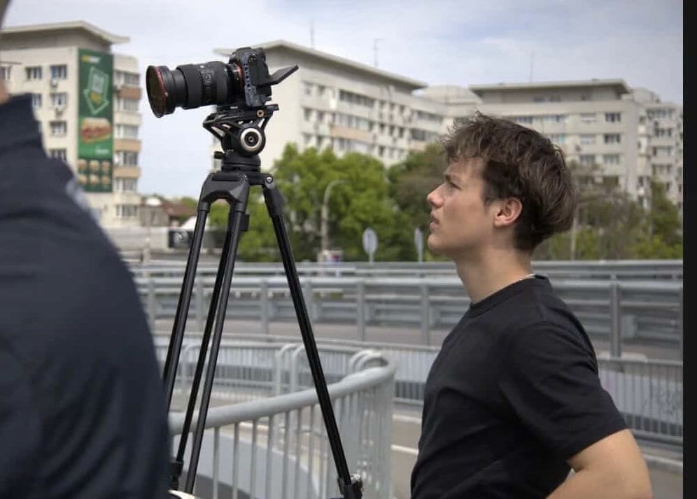 Alexandru Moise urmărind cadrul pe camera montată pe trepied, pe o pasarelă, cu blocuri în fundal.
